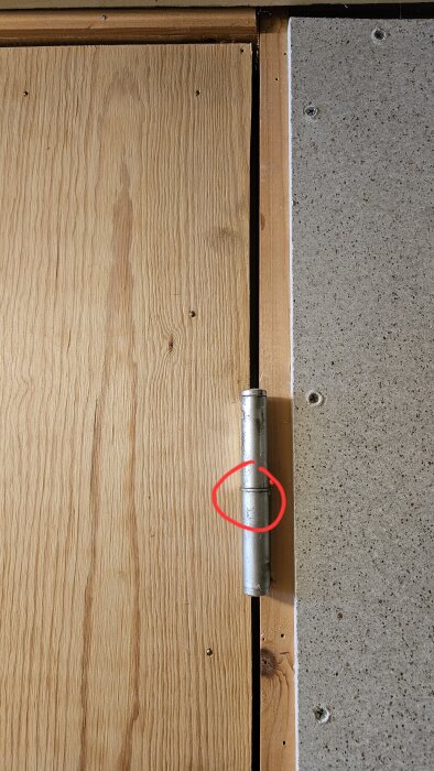 Trädörr med ett metallgångjärn markerat med en röd ring, bredvid en grå vägg.
