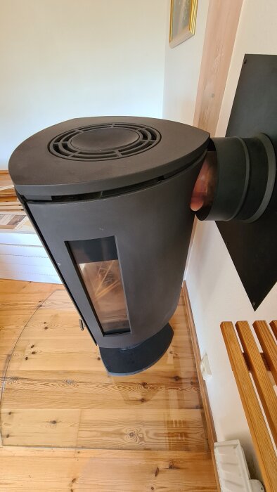 En svart Jötul F373-kamin med glasruta står på ett trägolv, delvis på en genomskinlig golvskiva. Rökröret är anslutet till väggen.