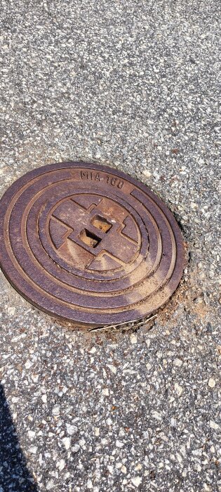 Rostigt brunnslock av metall på asfalterad yta med slitage och inskription MIA 160.