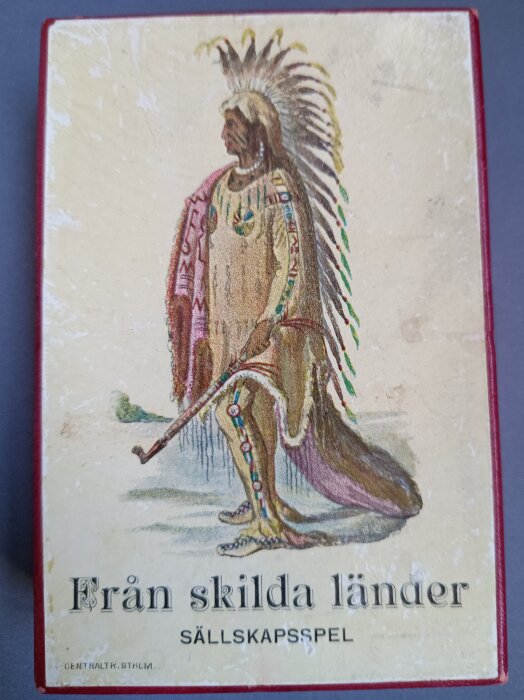 Omslag till sällskapsspelet "Från skilda länder" med illustration av person i traditionell klädsel och huvudbonad, ca 1900-talets början.