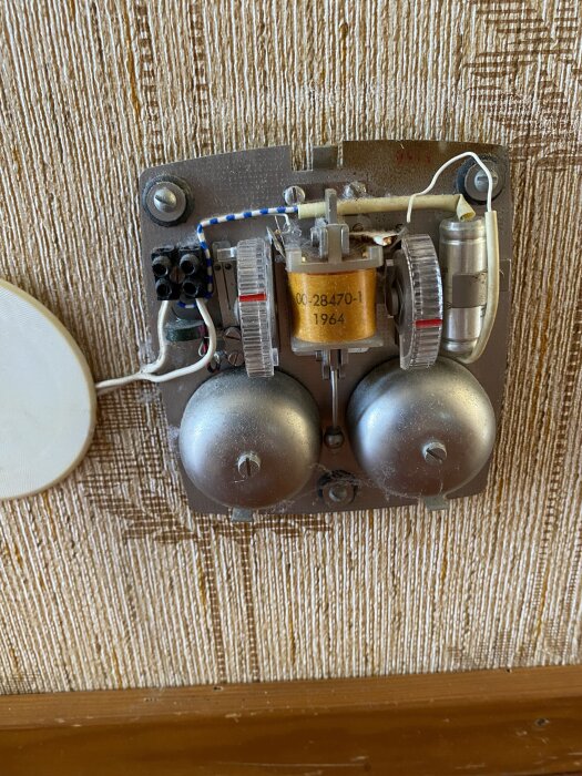 Ringklocka från 1960-talet monterad på tapetserad vägg i hall, med synliga kablar och mekanik.