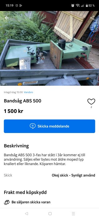 Bandsåg ABS 500 med grön färg står på släpvagn. En instruktionsbok syns ovanpå. En röd byggnad och växtlighet i bakgrunden.
