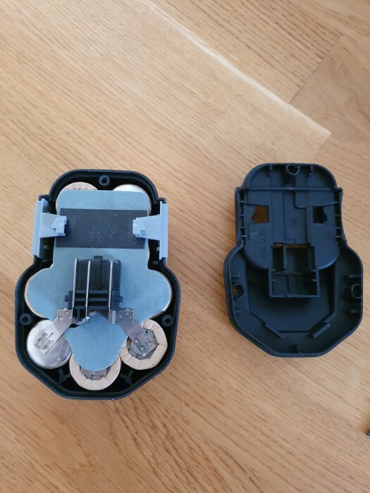 Öppet batteri med celler och kontakter synliga, batterilocket ligger bredvid det på ett trägolv.