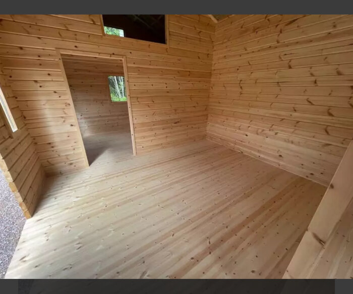 Interiör av en attefallstuga i trä med synliga bjälkar och öppet hål mot ett annat rum.