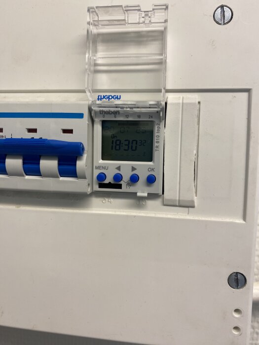 Timer på en elcentral med digital skärm visar tiden 18:30, omgiven av blå knappar och brytare i en vit ram.