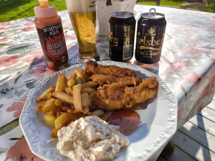 Tallrik med friterad kyckling, pommes frites och Dallassallad. En flaska Djävulssås, ölburkar och ett ölglas bredvid på en blommig duk.