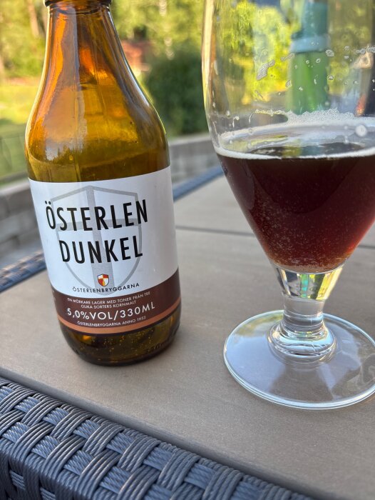 Ölflaska och glas med mörk öl på ett utebord i sommarmiljö, etikett "Österlen Dunkel" synlig.