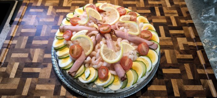Smörgåstårta dekorerad med räkor, citron, tomater, ägg, gurka och skinka på ett trämönstrat bord.