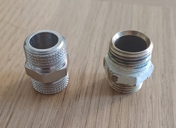 Två metallkopplingar placerade på ett trägolv, en ny och en gammal, med olika ytskick och gängor.
