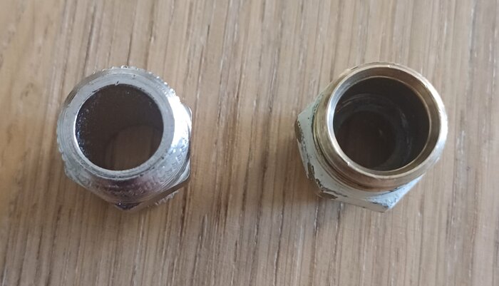 Två metallkopplingar för en kraninstallation, placerade på ett trägolv, där den vänstra kopplingen verkar större än den högra.