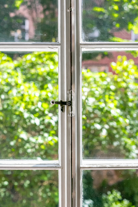 Gammaldags fönster med vågigt glas i 20-talslägenhet, grönska utanför.