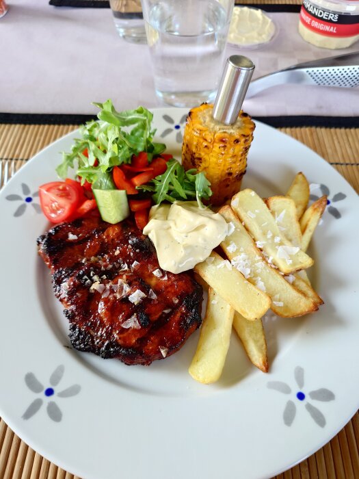 Grillad karré med pommes frites, bearnaisesås, majs, sallad, gurka, och tomat på en tallrik.