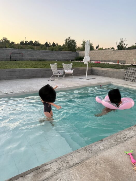 Utomhuspool med två barn som leker i vattnet. En rosa flytring syns, omgiven av en terrass och gräsmatta i skymningen.