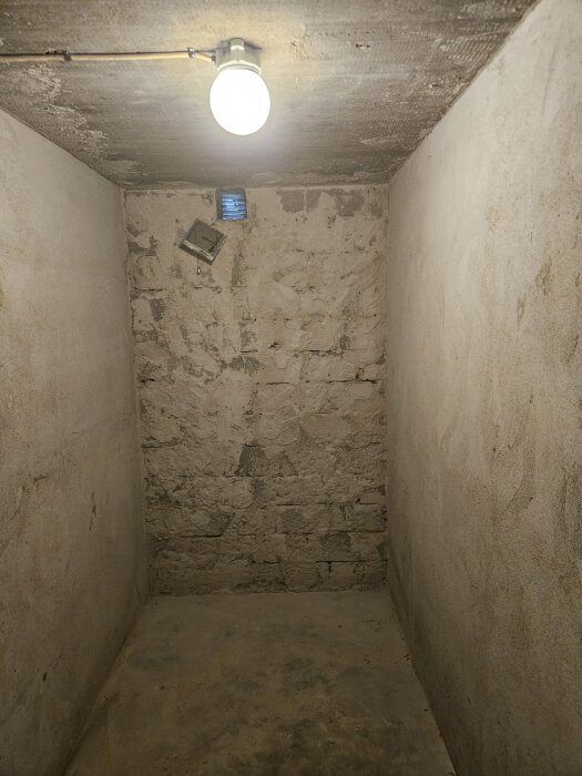 Källarvägg med borttagen träullsskiva, ojämn puts och slitet utseende, belyst av en enkel taklampa.