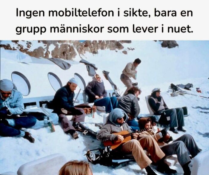 En grupp människor sitter och spelar gitarr på snön utanför ett hus med runda fönster. De ser avslappnade ut och njuter av solen.