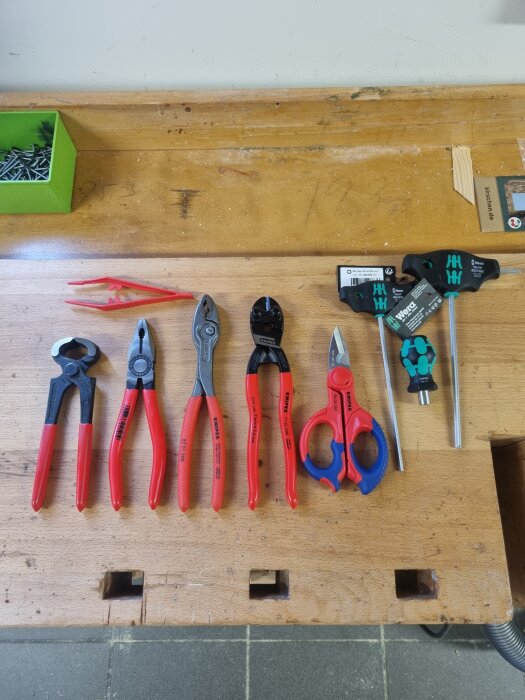 Röda och blå tänger och skruvmejslar på en träbänk bredvid grön låda med skruvar.