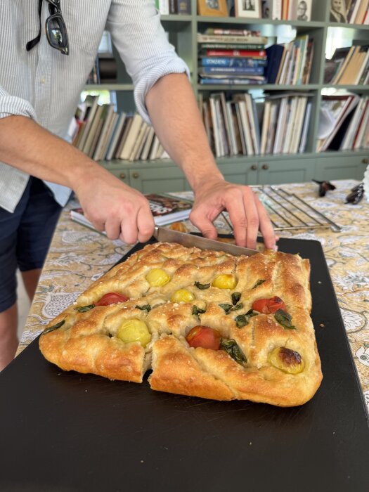 En person skär en hembakad focaccia med färgglada tomater och örter på en svart skärbräda, omgiven av en bokhylla i bakgrunden.