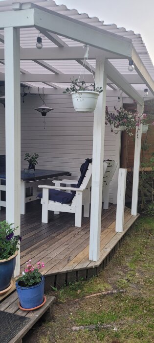 Utomhusveranda med tak, fyra stolpar och en stolpe i mitten, omgiven av växter i krukor, utan staket.