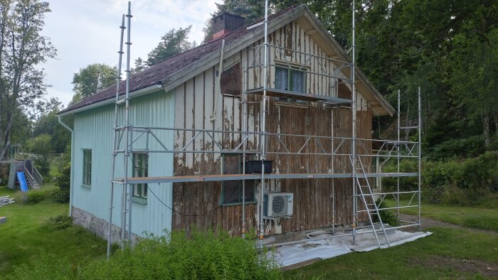 Husrenovering med byggställning, vägg skrapas trärent. Delvis målad fasad. Värmepump synlig, omgiven av grönska och träd.
