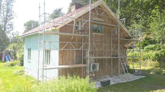 Stående rödbrun stuga med ställningar för målning och renovering. Delvis färdigmålad, luftvärmepump synlig, omgiven av grönska.