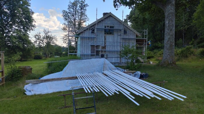 Utsikt över en stuga med byggställning under renovering, vitmålade trälister ligger på gräset under en presenning.