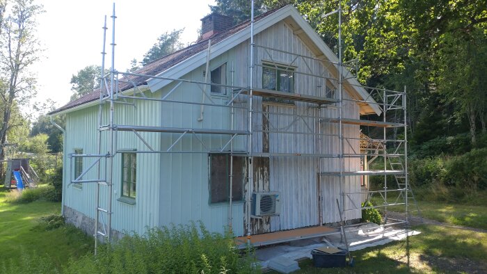 Stuga under renovering med byggställning; vägg målad med grundfärg, innehåller linoljefärginstruktioner, arbetsutrustning och en värmepump.