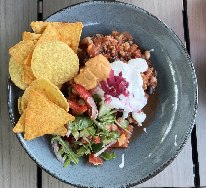 Chili con carne med nachos, sallad, ostsås, picklad rödlök och gräddfil serverad på en tallrik.