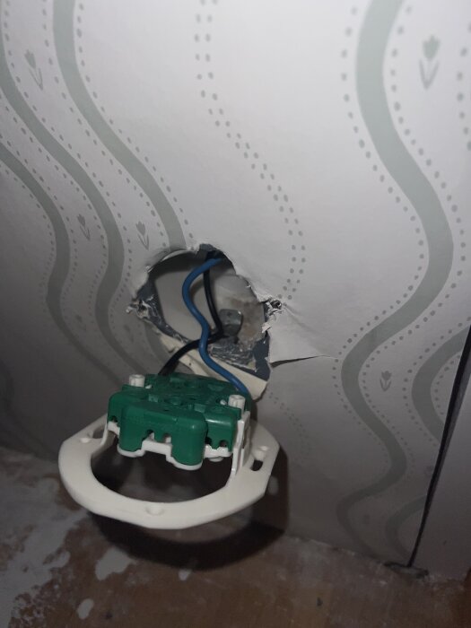 Eluttag med grönt och vitt montage hänger löst ur hål i väggen med blommigt tapetmönster, metallring försvårar installation.