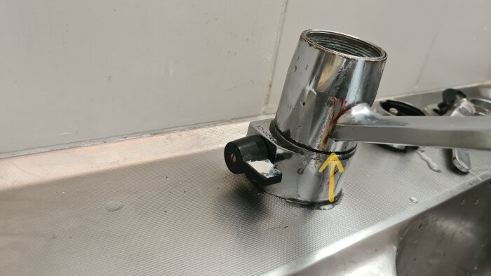 Kromad blandare med lossat handtag och en gul pil markerar möjlig plats att bända för access till gummipackningen. Skruv synlig i spegelbilden.