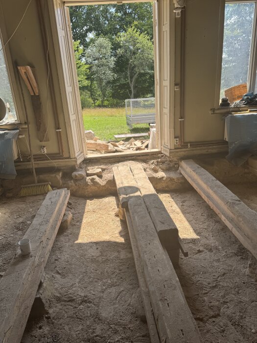 Öppet dörrparti med vy mot trädgård; inomhus golvuppriven med exponerade bjälkar och jordgolv i gammalt stenhus under renovering.