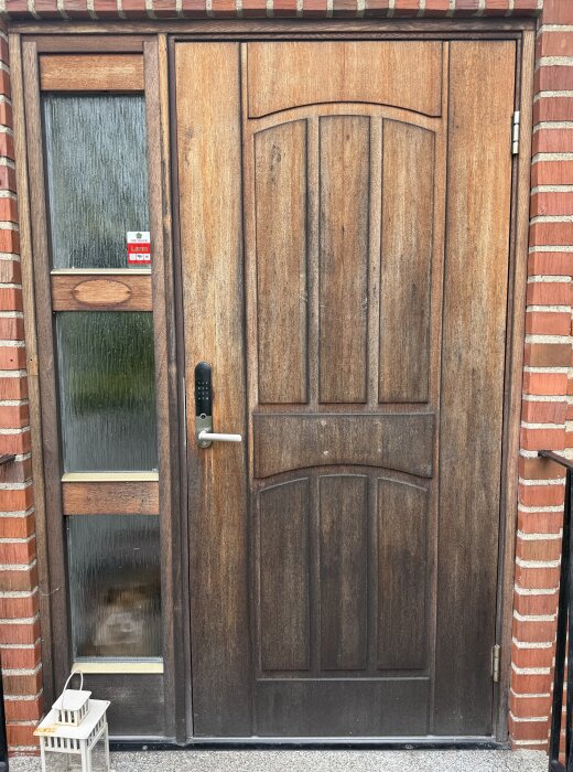 Solblekt och matt ytterdörr i trä med glasparti bredvid, rutorna har ornamenterat glas. Dörren har en modern låssats och en lykta står framför.