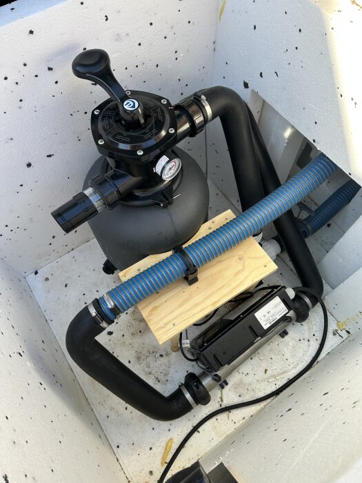 Poolfilter med svart handtag och blå slang, placerat i ett hörn av en poolpumpinstallation med träunderstöd och rörkopplingar.