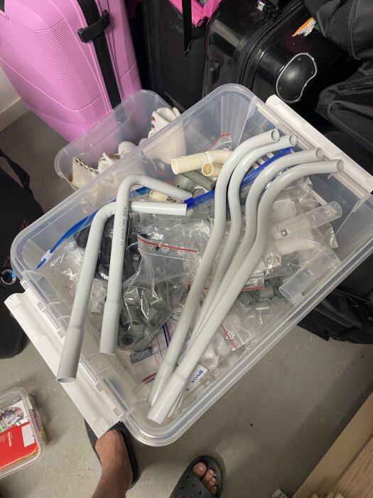 Plastback med olika rörböjar och kopplingar i ett förråd, omgiven av resväskor.