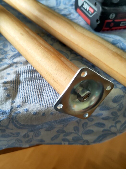 Stolsben av trä med förstärkning av en mutter vid metallinfästning, liggande på en blåmönstrad duk. Bakgrund: en skruvdragare.