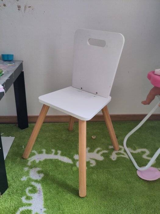 Barnskrivbordsstol med vita sits och ryggstöd, träben, står på en grön matta; infästningsreparation med mutter har gjorts.