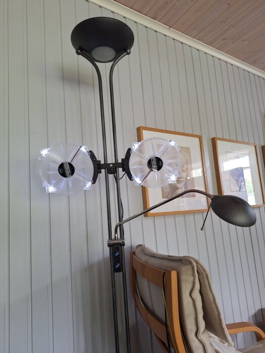 Två datorfläktar monterade på en stående golvlampa bredvid en fåtölj, i ett rum med panelvägg och tavlor.