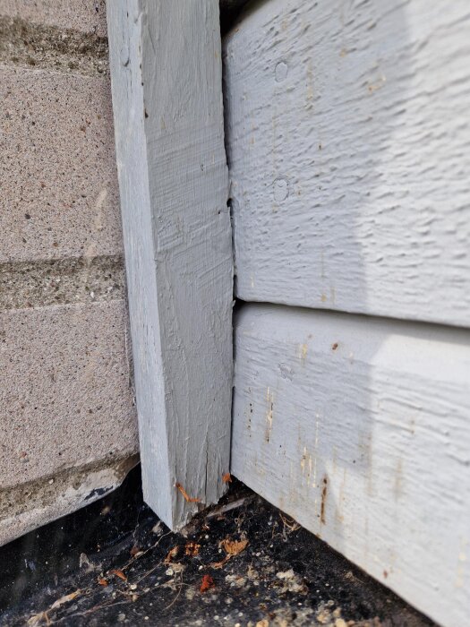 Slitet gråmålat fönsterfoder med färgfläckar och smuts vid murad vägg, träet är sprucket i hörnet.