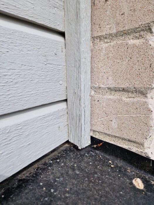 Fönsterfoder och fasad med avskavd färg vid tegelvägg; träet ser slitet ut och ligger ovanpå ett smutsigt underlag.