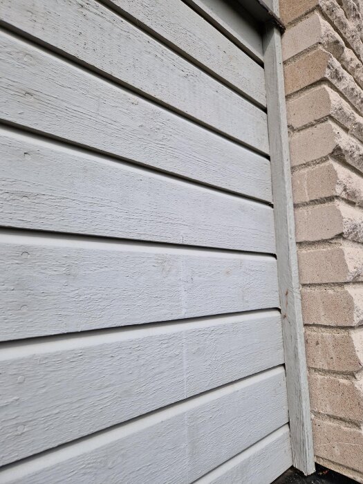 Fasad av träpanel i behov av underhåll, med intilliggande tegelvägg. Fönsterfoder och målning diskuteras för framtida renovering.