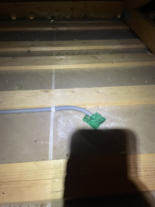 Böjd kabel fastsatt med klammer på golv av trä och gips, med grönt markeringsband intill och skugga i förgrunden.