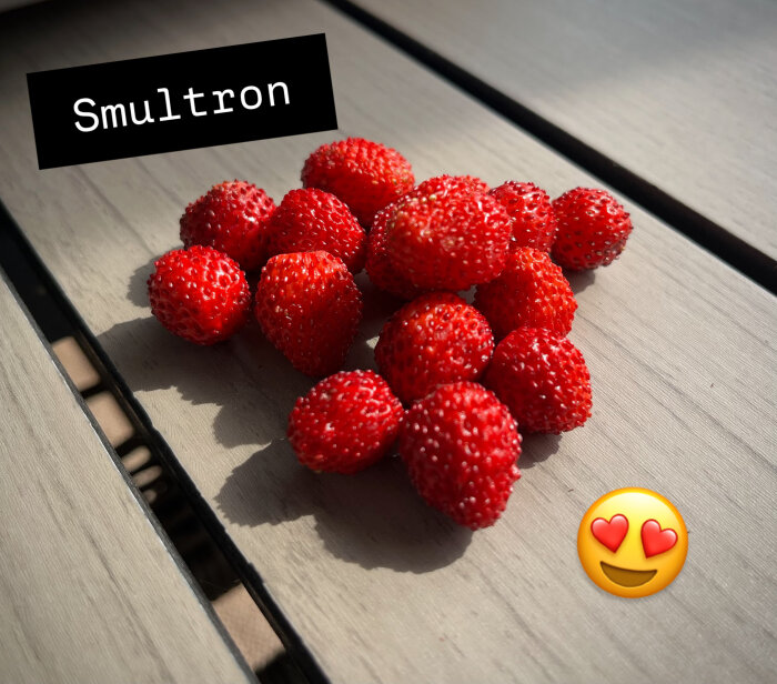 Färska smultron ligger på ett träbord i solljus, med texten "Smultron" och en emoji med hjärtformade ögon bredvid.