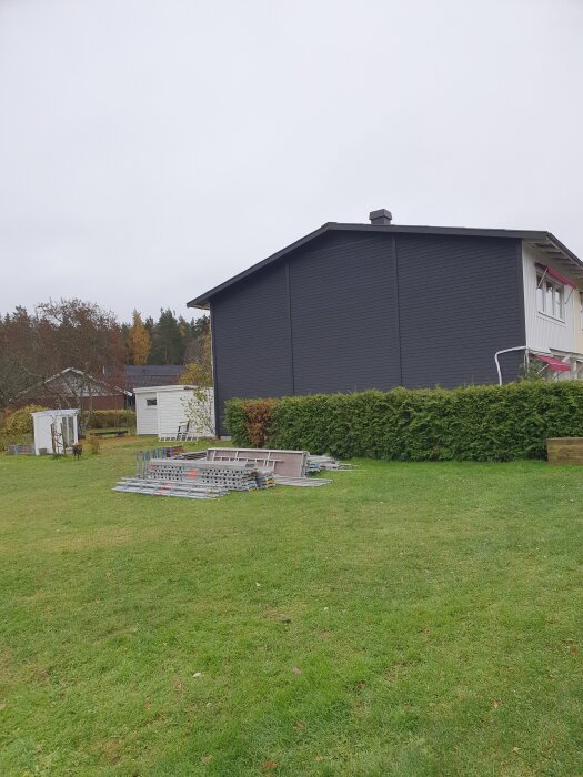 En hög med byggnadsställningar ligger på en gräsmatta bredvid ett mörkgrått hus och en häck.