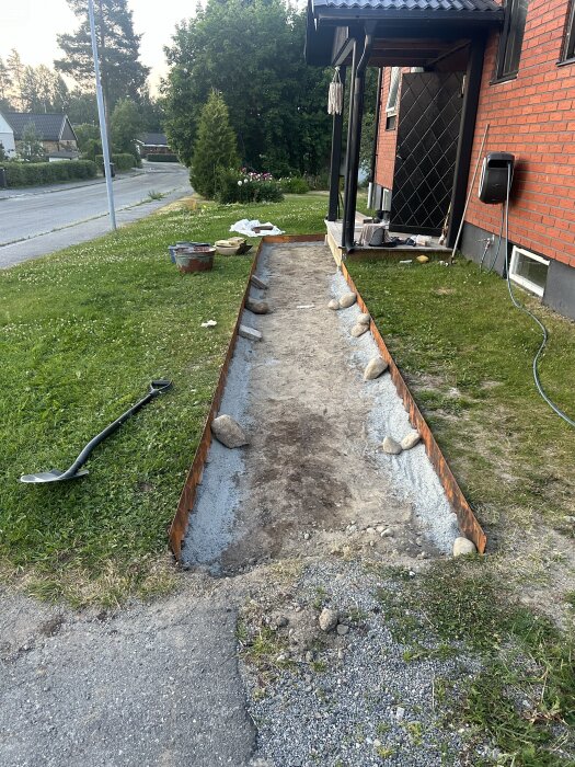 Gångstig under konstruktion vid entré, med cortenplåtskanter och stenar längs sidorna. Verktyg och material synliga i bakgrunden.