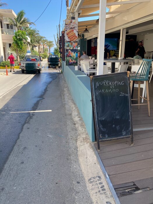 Skylt utanför restaurang med text om match Norrköping vs Värnamo kl 20:00, gata med bilar och palmer i bakgrunden under klar himmel.