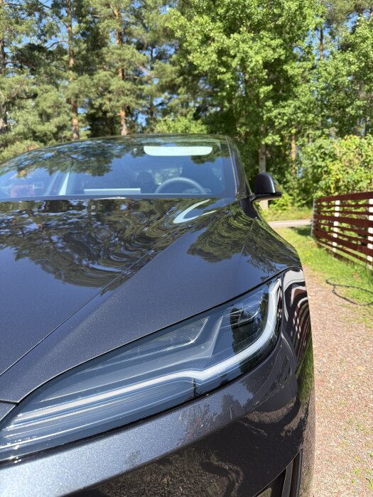 Svart Tesla Model 3 2024, nyputsad med stark glans. Står på en uppfart omgiven av gröna träd och en röd staketstruktur.