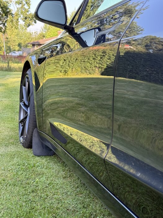 Svart bil (Model 3 2024) med hög glans reflekterar omgivningen, stående på en gräsmatta, resultat av nyligen utförd polering och lackskydd.