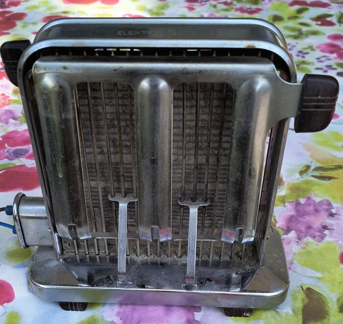 Gammaldags brödrost med metallarmar för att vända brödskivor automatiskt, stående på ett blommigt bord.