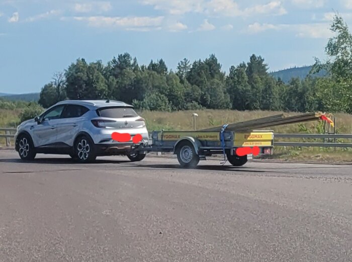 Bil med släpkärra fylld med långa träbalkar på en landsväg, omgiven av skog och landskap i bakgrunden.