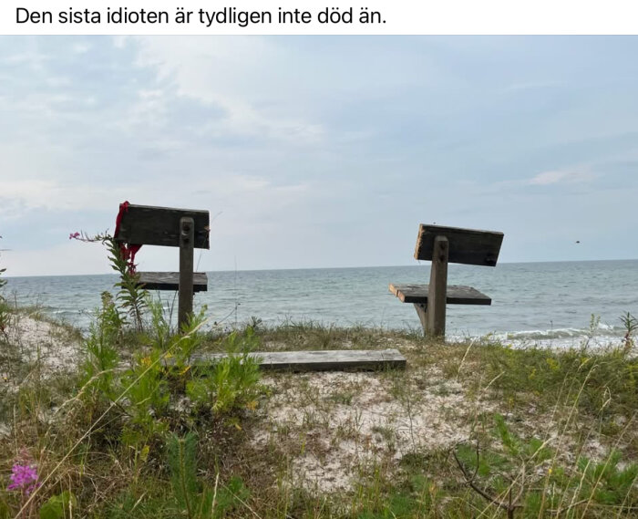 Strand med havsutsikt, två hemgjorda trästolar med ryggstöd i olika riktningar, sandig mark med vegetation, mulen himmel.