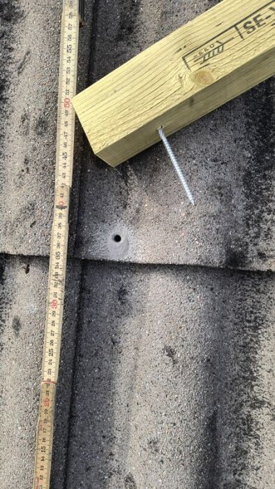 Regelbit av tryckat virke med skruv fast i takpanna, bredvid måttstock och borrat hål, förberedelser för solpanelinstallation.